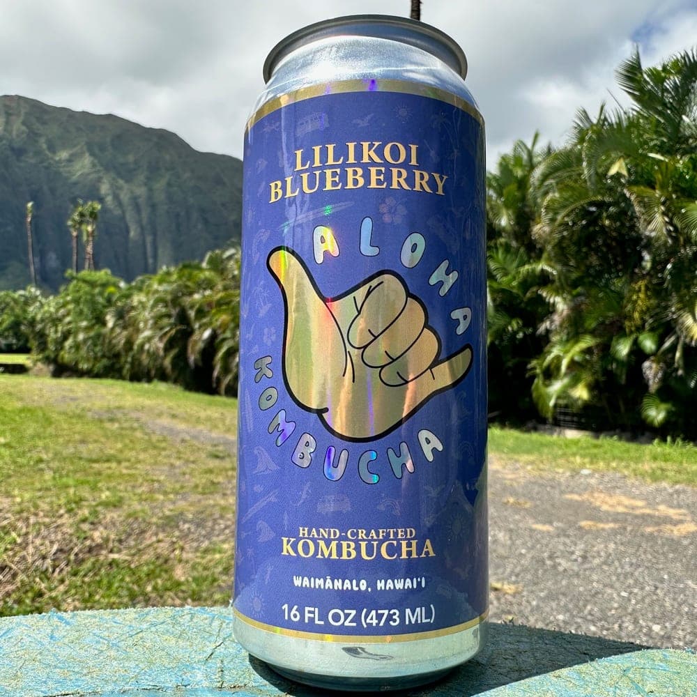A Lilikoi Blueberry Aloha Kombucha can with the Koʻolau mountains and palms behind it